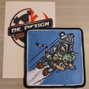 Könnte beinhalten: Gestickter Patch mit blauem Hintergrund, auf dem ein weißer und grauer Mandalorianer mit einem grünen Grogu-Charakter fliegt. Der Mandalorianer trägt einen Helm und hält ein Blastergewehr. Der Text "DK DESIGN" ist im Hintergrund sichtbar.
