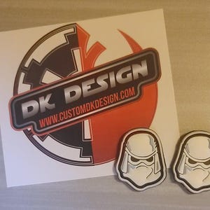 Könnte beinhalten: Ein weißer Aufkleber mit einem schwarzen und roten Star Wars Rebel Alliance-Symbol und dem Text "DK DESIGN www.customdkdesign.com". Zwei weiße, plastische Aufkleber in Form von Stormtrooper-Helmen befinden sich unterhalb des größeren Aufklebers.