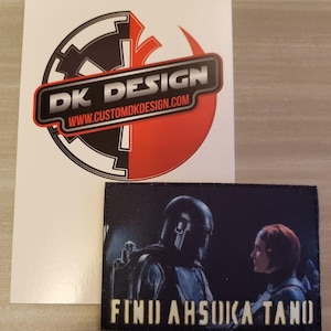 May include: A black and red sticker with the text "DK DESIGN" and the website "www.customdkdesign.com". Below the sticker is a black patch with a blue and white image of two figures, one in a helmet and the other with red hair. The text "FIND AHSOKA TANO" is printed in yellow on the patch.