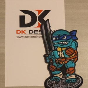 Puede incluir: Un parche bordado de una Tortuga Ninja Adolescente sosteniendo una katana. La tortuga es azul, verde y naranja. El parche está sobre un fondo gris con un diseño de rejilla de alcantarilla.
