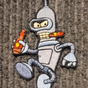 Può includere: Una toppa ricamata di un robot che tiene una bottiglia e fuma un sigaro. Il robot è blu e grigio e ha un'espressione severa.