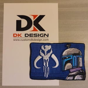 Pode incluir: Patch bordado azul e branco com um capacete mandaloriano e o símbolo do clã mandaloriano. O patch está sobre um fundo branco com um cartão de visita que diz "DK DESIGN" e "www.customdkdesign.com".