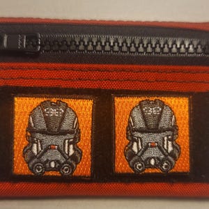 Könnte beinhalten: Ein rechteckiges rotes und schwarzes Portemonnaie mit einem schwarzen Reißverschluss. Zwei orangefarbene Quadrate mit gestickten grauen und weißen Helmdesigns befinden sich auf der Vorderseite. Eine kleine schwarze Stoffschlaufe ist am Reißverschluss befestigt.
