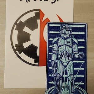 May include: A blue and white embroidered patch of a Star Wars clone trooper standing in front of a blue and white striped background. The patch has the text "Dk Design" on the top left corner.
