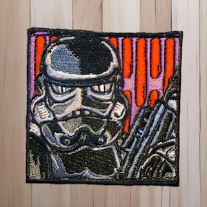 以下が含まれることがあります： 黒、グレー、白のストームトルーパーのヘルメットが刺繍されたパッチ。背景にはオレンジと紫の縦縞があります。パッチは正方形で、黒い縁取りがあります。