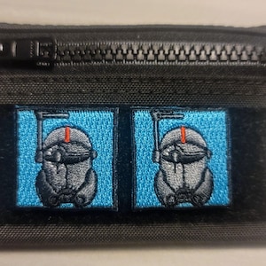 May include: A black zippered pouch with two embroidered patches. Each patch features a blue background with a white and gray robot design. The robot has a red antenna and a black visor.