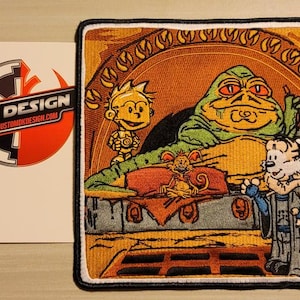 May include: A colorful embroidered patch featuring a green Jabba the Hutt-like character with a yellow and orange creature and a tiger-like character in a blue robe. The text "DK DESIGN" is visible in the background.