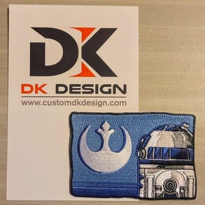 May include: A blue and white embroidered patch featuring a stylized R2-D2 droid and a white Rebel Alliance symbol. The patch is rectangular with a dark blue border. A white card with the text "DK DESIGN" and website address is in the background.