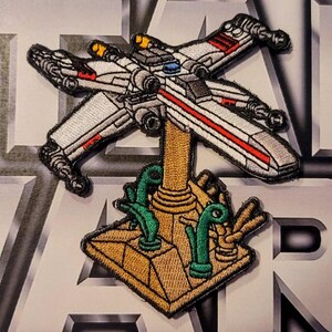 Może przedstawiać: Naszywkę z haftowanym białym i czerwonym myśliwcem X-Wing z serii Star Wars. Myśliwiec jest umieszczony na brązowym stojaku z zielonymi roślinami rosnącymi wokół niego.