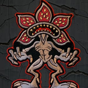May include: Embroidered patch featuring a Demogorgon monster design. The patch has a red border, with the monster in shades of tan, black, and red. The monster has a flower-like head with sharp teeth and clawed hands and feet.
