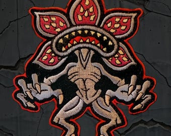 Pode incluir: Patch bordado com um design de monstro Demogorgon. O patch tem uma borda vermelha, com o monstro em tons de bege, preto e vermelho. O monstro tem uma cabeça em forma de flor com dentes afiados e mãos e pés com garras.