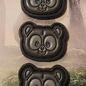 Może przedstawiać: Trzy czarne i szare łatki z postacią o uśmiechniętej twarzy i dużymi oczami. Łatki są okrągłe i mają czarną obwódkę.