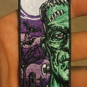 May include: A rectangular embroidered patch featuring a green monster face on the right side and a purple graveyard scene with a full moon, bare trees, tombstones, and flying bats on the left side, all enclosed by a black stitched border.
