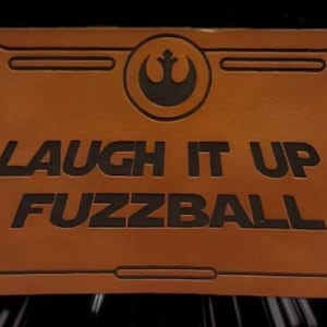 May include: A rectangular, brown plaque with the text "LAUGH IT UP, FUZZBALL" in black. The plaque features a black Rebel Alliance symbol and a decorative border. The background is a dark space scene with white streaks.