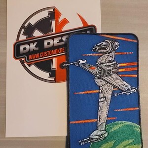 May include: A blue and green embroidered patch featuring a white spaceship firing orange lasers against a blue background. The spaceship is flying over a green planet. The patch has the text "DK DESIGNS" and "WWW.CUSTOMDKDESIGNS.COM" on the bottom.