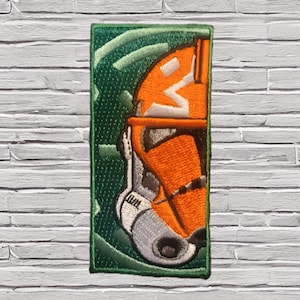 May include: A rectangular embroidered patch featuring a profile view of a white and orange helmet with a dark visor. The background is a deep green with abstract swirling lines. The design is enclosed within a thin green stitched border.