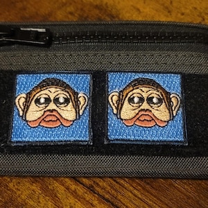 May include: A dark grey zipped pouch with two embroidered patches. Each patch features a cartoon monkey face on a blue square. The monkey has large eyes, a red mouth, and tan fur. The pouch has a black zip and a small black loop.
