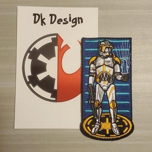 Puede incluir: Un parche bordado azul y amarillo con un clon de Star Wars con un rifle bláster. El clon está de pie sobre un símbolo circular amarillo y negro. El texto "Dk Design" es visible en una tarjeta blanca detrás del parche.