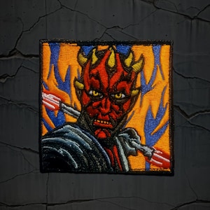 May include: Embroidered patch featuring a detailed portrait of Darth Maul from Star Wars. The patch showcases Maul's red and black face with yellow eyes, set against an orange and blue background. The design includes his double-bladed lightsaber.