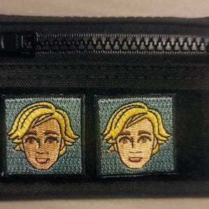 May include: Black zippered pouch with two embroidered patches. Each patch features a blonde-haired person's face with a smiling expression. The patches are square and set against a teal background. The pouch has a black zipper and a small fabric loop.