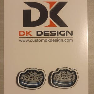 May include: Two small, white, gray, and blue Millennium Falcon spaceship-shaped pins. The pins are on a white background with the text "DK DESIGN" and "www.customdkdesign.com" below.