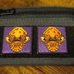 May include: A black zippered pouch with two embroidered patches. The patches are purple with orange alien-like creatures with red accents.