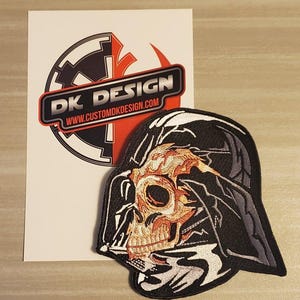 Puede incluir: Un parche bordado en blanco y negro de una calavera con un casco de Darth Vader. El parche está sobre un fondo blanco con una pegatina de un logotipo rojo y negro que dice "DK DESIGN" y "www.customdkdesign.com".