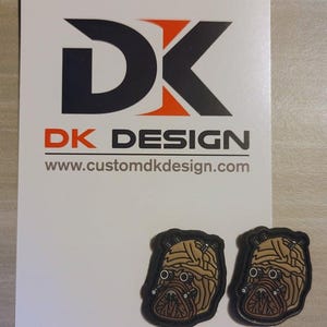 May include: Two brown and black rubber patches with a detailed image of a creature with a large head and multiple eyes. The patches are on a white background with the text "DK DESIGN" and "www.customdkdesign.com".