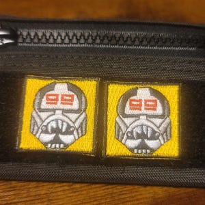 May include: Black zippered pouch with two yellow patches featuring embroidered white and red stormtrooper helmets with the number "99" on each.