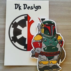 May include: A white card with the text "Dk Design" and a black and white image of the Star Wars Imperial symbol. Next to the card is a green, red, and yellow embroidered patch of a Star Wars Boba Fett character.
