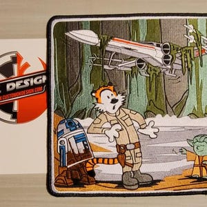 Op de afbeelding: Een geborduurde patch met een cartoon-tijger die een pilotenuniform draagt, naast R2-D2 en Yoda. De tijger kijkt naar rechts, met een verraste uitdrukking. De achtergrond is een groen bos met een witte X-Wing-jager die erboven vliegt. De patch heeft een zwarte rand en de tekst "DK DESIGN www.customdkdesign.com" is afgedrukt in de linkerbenedenhoek.