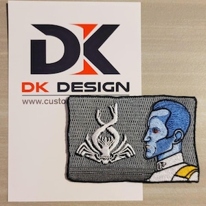 May include: A gray embroidered patch with a white alien creature and a blue-skinned humanoid figure with a white and yellow uniform. The patch has the text "DK DESIGN" on the bottom.