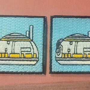 Puede incluir: Dos parches bordados con una nave espacial blanca, amarilla y gris sobre un fondo azul claro.