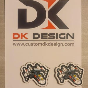 Könnte beinhalten: Zwei weiße, rote und gelbe X-Wing-Sternenjäger-Patches mit schwarzen Details. Die Patches befinden sich auf einem weißen Hintergrund mit dem Text "DK DESIGN" und "www.customdkdesign.com" darunter.