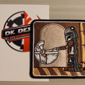 May include: Embroidered patch featuring a cartoon Mandalorian character holding a baby Yoda in a crib. The patch has a brown, tan, and black color scheme. The text "DK DES" and "WWW.CUSTOMDKDES" is visible in the background.