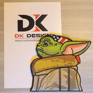 Puede incluir: Parche bordado con un personaje verde de orejas grandes que lleva un pañuelo rojo, blanco y azul. El personaje está sentado en una prenda marrón con capucha. El parche tiene el texto "DK DESIGN" y "www.customdkdesign.com".