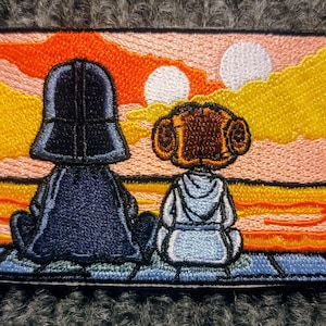 Puede incluir: Parche bordado con Darth Vader y la Princesa Leia, de pie uno al lado del otro, mirando un atardecer con dos soles. El parche está sobre un fondo rosa y naranja con nubes amarillas.