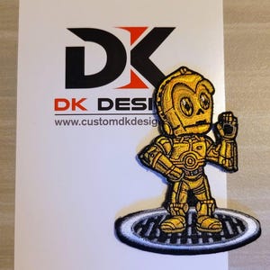 May include: A yellow embroidered patch of a cartoon C-3PO character from Star Wars. The patch is on a white background with the text "DK DESI" and "www.customdkdesign.com".