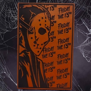 May include: Orange and black rectangular sign featuring a stylized illustration of a hockey mask from the movie "Friday the 13th." The words "Friday the 13th" are repeated multiple times on the orange background. The sign is framed in black.