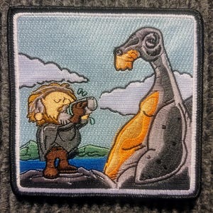 May include: Embroidered patch featuring a cartoon of a small, bearded man in a brown coat drinking from a cup, while a large, gray, orange, and black creature with horns looks on.