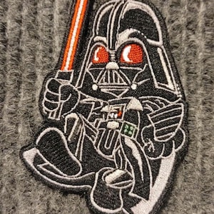 Puede incluir: Un parche bordado en blanco y negro de Darth Vader sosteniendo un sable de luz rojo. El parche está sobre una tela de punto gris.