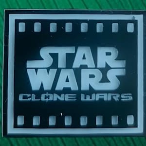 Może przedstawiać: Kwadratowa czarno-biała przypinka z logo Star Wars Clone Wars pośrodku. Wzór jest otoczony białą ramką z motywem taśmy filmowej na górnej i dolnej krawędzi, na której wyraźnie widoczny jest napis Star Wars Clone Wars.