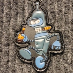 Puede incluir: Un parche con un personaje de robot de dibujos animados en tonos azules, sosteniendo una botella y un cigarro. El robot tiene una expresión severa y está delineado en negro. El parche está sobre un fondo gris texturizado.