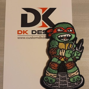 May include: An embroidered patch featuring a cartoon Teenage Mutant Ninja Turtle with a red bandana, green skin, and a yellow undershirt. The turtle is holding a pair of sai and has an angry expression. The patch is on a white background with the text "DK DESIGNS" and a website address.