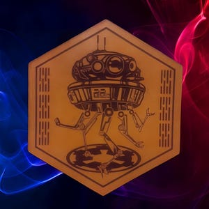 May include: A tan, hexagonal plaque with a black illustration of a Star Wars Imperial probe droid. The droid is in the centre, with the Imperial symbol below. The background has blue and red smoke effects.