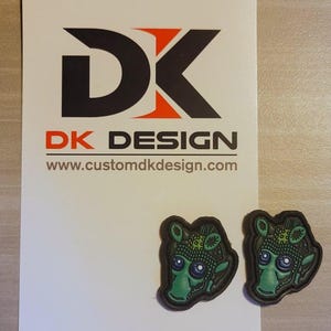 May include: Two green and brown rubber patches featuring a cartoon alien face. The patches are on a white card with the text "DK DESIGN" and "www.customdkdesign.com".