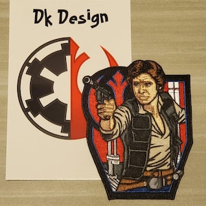 Puede incluir: Parche bordado con Han Solo de Star Wars sosteniendo un bláster. El parche es un rectángulo azul oscuro con un símbolo de la Alianza Rebelde rojo y azul en el fondo. El texto "Dk Design" es visible en una tarjeta blanca detrás del parche.
