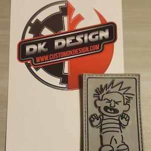 Könnte beinhalten: Ein grauer Patch mit schwarzem Umriss und weißem, genähtem Rand. Der Patch zeigt eine Schwarzweißillustration eines Cartoon-Jungen mit stacheligem Haar, der ein gestreiftes Hemd und eine Hose trägt und einen verzweifelten Ausdruck hat. Der Text "DK DESIGN" und "WWW.CUSTOMDKDESIGN.COM" ist auf einem weißen Aufkleber mit rotem und schwarzem Hintergrund gedruckt.