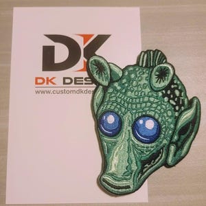 May include: Green embroidered patch of a fictional alien character with large blue eyes. The patch is on a white background with the text "DK DESIGNS" and "www.customdkdesigns.com".