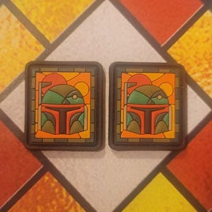 May include: Two square-shaped patches with a stained glass design featuring a green, red, and brown helmet with a yellow background. The patches are made of a textured material and have a black border.